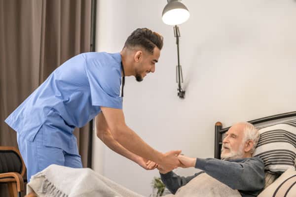 A caregiver helps a senior man reposition in bed to prevent bed sores.