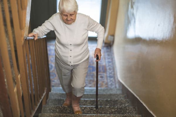 An older woman walks carefully up the stairs with her cane, highlighting the need to assess the safety of an older loved one’s home.